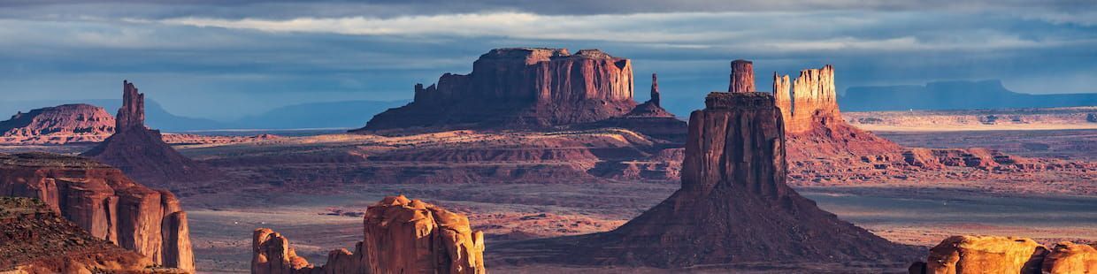 Panoramic view of Monument Valley in Arizona, showcasing towering red sandstone buttes and mesas under a cloudy sky. The vibrant colors and dramatic landscape capture the unique beauty of this iconic desert scene.