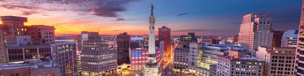 Aerial view of a cityscape at sunset in Indiana, featuring a central monument surrounded by tall buildings. The sky is a vibrant blend of orange, pink, and purple hues, casting a warm glow over the urban landscape—much like the bright future offered by online education master's degree programs.