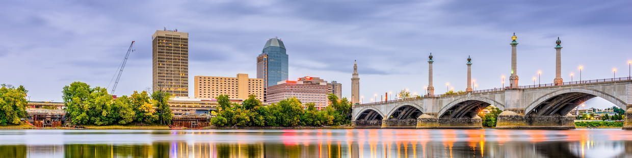 A city skyline featuring a tall building and a bridge over a calm river in Massachusetts, where cloudy skies and bridge lights reflect on the water. Trees along the riverbank add a touch of greenery to the scene, echoing the serenity ideal for pursuing an Online Master's in Education.