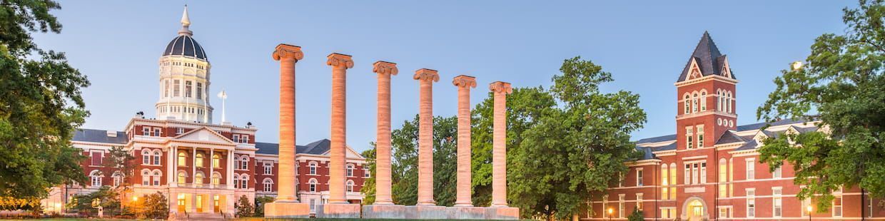 A historic university campus in Arizona, featuring a domed building, six tall stone columns, and a brick structure with a pointed roof, all nestled among lush green trees under a clear blue sky. The perfect setting for pursuing an online master’s in education or exploring various education programs.