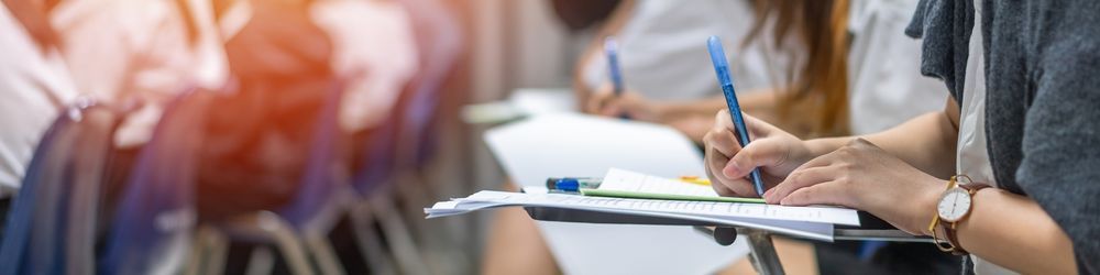 Students sitting in a classroom take notes with pens on notepads. They are seated in rows with writing surfaces, and the scene is brightly lit.